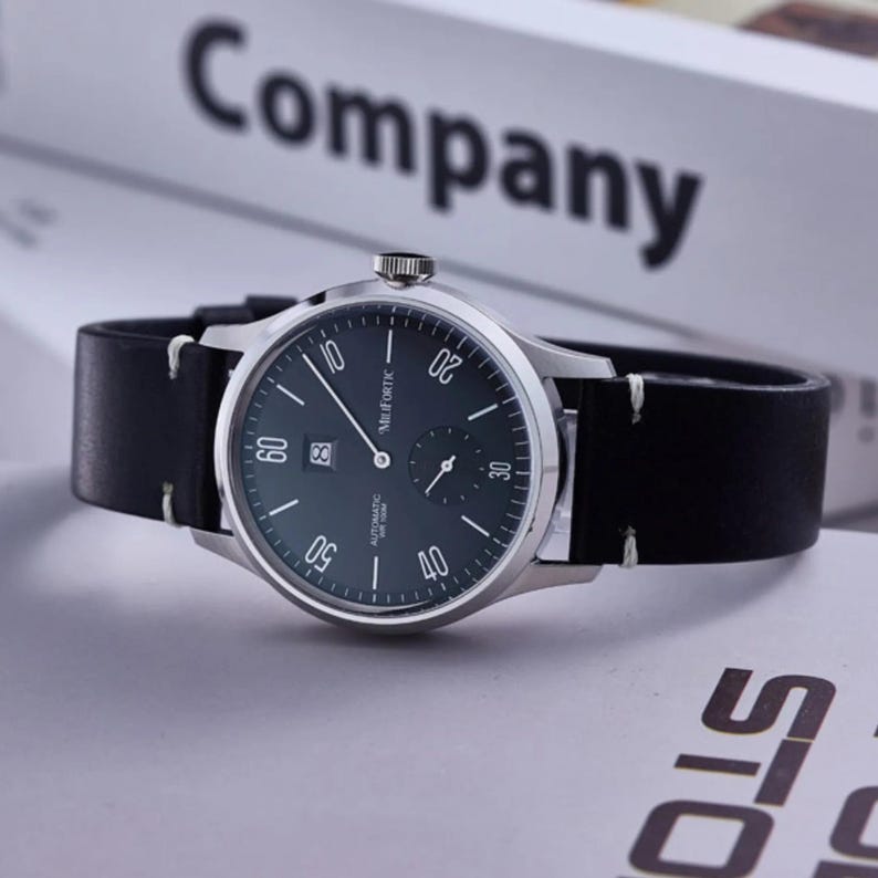 May include: A silver-toned wristwatch with a black leather band. The watch face is dark green with white numerals and hands. The watch has a date display and the text "MuFORTIC" is visible. The word "Company" is in the background.