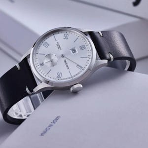 May include: Silver-toned wristwatch with a white face, black leather band, and a date display. The watch features a small seconds dial and the brand name "Muifortc". The band is stitched with white thread.