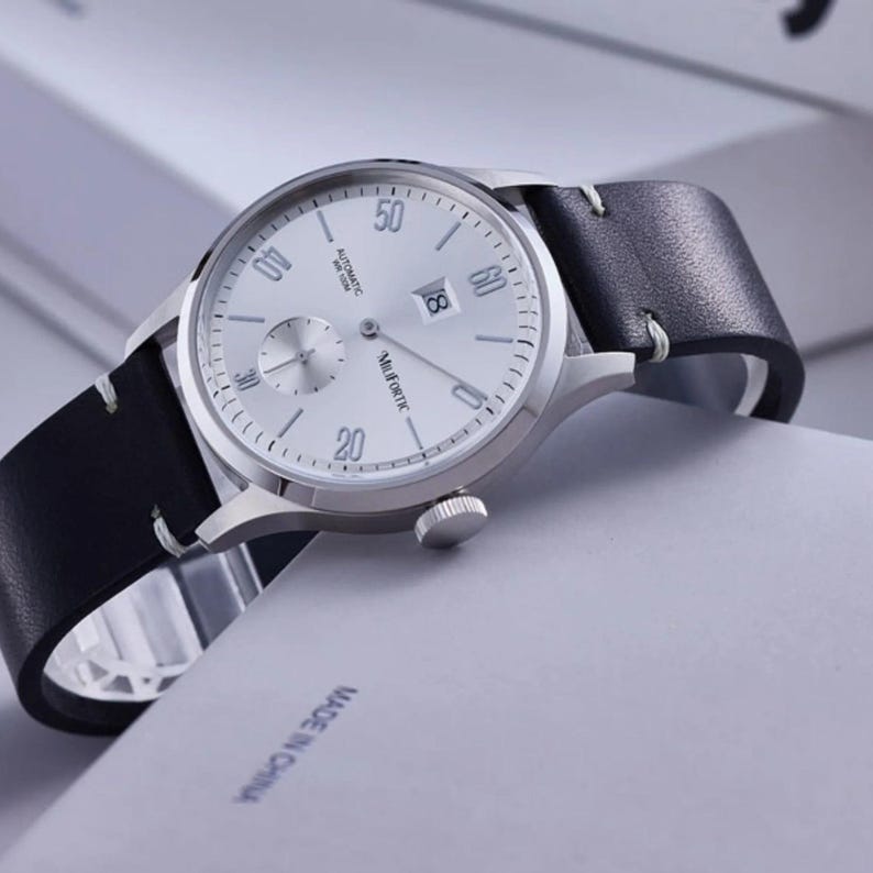 May include: Silver-toned wristwatch with a white face, black leather band, and a date display. The watch features a small seconds dial and the brand name "Muifortc". The band is stitched with white thread.