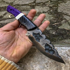 Op de afbeelding: Een op maat gemaakt mes met een paarse handgreep, een witte spacer en een koolstofvezel grip. Het lemmet heeft een uniek, getextureerd ontwerp met een donkere afwerking. Het mes wordt in een hand gehouden, wat het vakmanschap benadrukt.