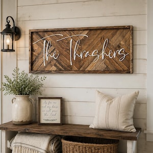 May include: A rectangular wooden wall sign featuring a dark brown herringbone pattern background and white 3D lettering that reads The Thrashers. The sign is mounted horizontally on a white horizontal plank wall above a wooden bench.