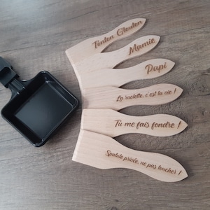 Op de afbeelding: Een set van vijf houten spatels voor raclette, elk met een andere inscriptie. De spatels zijn in een waaier vorm gerangschikt, met de inscriptie "Tonton Glouton" bovenaan en "Spatule privée, ne pas toucher!" onderaan. De spatels zijn gemaakt van lichtbruin hout en hebben een natuurlijke afwerking.
