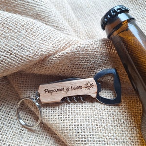 Peut inclure: Un ouvre-bouteille en bois avec un tire-bouchon et un petit couteau. Le porte-clés a le texte "Papounet je t'aime" gravé dessus. Le porte-clés est sur un fond en toile de jute avec une bouteille en verre brun.