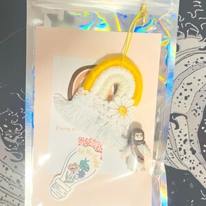 Puede incluir: Una bolsa de plástico transparente que contiene un macramé de arcoíris amarillo, blanco y marrón, una pequeña figura de Jesús y una tarjeta con el texto "Enjoy your..." y "Be the light" con un gráfico de bombilla.