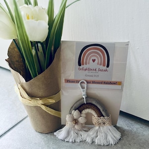 Puede incluir: Un llavero de arcoíris hecho a mano con una combinación de colores gris, marrón y crema, adornado con un adorno floral y flecos blancos. El llavero está adjunto a una tarjeta con el texto "Enlightened Sarah Art Boutique" y "Please Enjoy your Blessed Rainbow!"