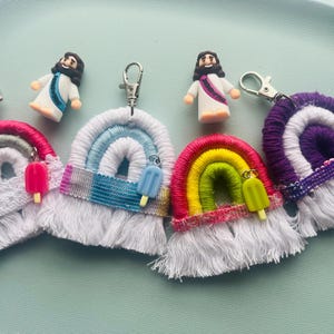 Puede incluir: Cuatro llaveros de arcoíris de macramé con cierres de mosquetón plateados. Cada uno presenta un esquema de color único, flecos blancos y un pequeño dije de paleta. Dos pequeñas figuras de una persona con túnica blanca entre los llaveros.