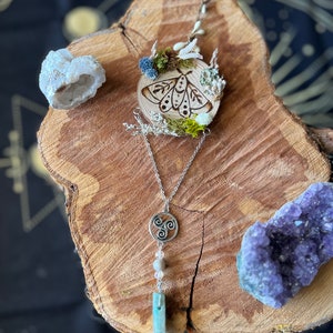 May include: A silver necklace with a triquetra pendant, a light blue crystal, and a white bead. The necklace is displayed on a wooden surface with a small wooden circle featuring a burned image of a moth with white flowers and moss.