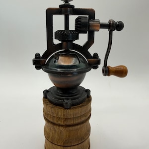 May include: A traditional pepper mill with a dark bronze finish and a wooden base. The mill has a hand crank and a decorative top. The design includes a glass or dark-coloured container for the peppercorns. The overall aesthetic is rustic and classic, reminiscent of older designs.