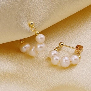 May include: A pair of gold hoop earrings with small white pearls. The earrings are on a light beige fabric background.