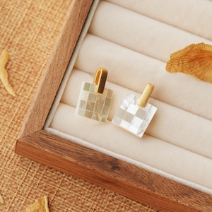 May include: Two square-shaped earrings with a gold-tone metal setting and a white mother-of-pearl inlay. The earrings are displayed in a wooden box with a white velvet lining.