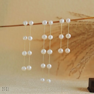 May include: Delicate pearl drop earrings. Each earring showcases a sequence of white, round pearls hanging from a fine gold chain. The earrings are presented against a soft, neutral backdrop, accentuating their classic design and sophisticated charm. A timeless accessory.