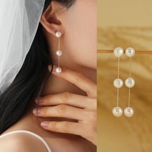 May include: Gold-tone drop earrings with three white pearls hanging from fine chains. The earrings are displayed on a person's ear and also shown separately. A classic jewellery item, suitable for special occasions or everyday wear.