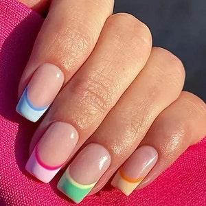 Pode incluir: Close-up de uma mão com unhas quadradas. Cada unha apresenta uma ponta francesa de cor pastel diferente: azul claro, rosa, verde e laranja. As unhas têm um acabamento brilhante e a pele é clara. O fundo é um tecido rosa brilhante.