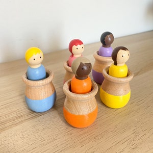 Puede incluir: Seis muñecas de madera con clavijas, cada una con un color de pelo y un tono de piel diferentes, están sentadas dentro de macetas de madera pintadas. Las macetas están pintadas en diferentes colores, incluyendo azul, amarillo, naranja y morado.