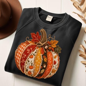 Peut inclure: Un sweat-shirt noir orné d'un grand motif central de citrouille composé de divers empiècements en tissu orange, jaune et blanc avec des motifs à carreaux, floraux et de feuilles. Le design inclut des bords cousus, des glands et des feuilles d'automne.