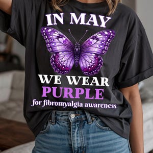 May include: Dark gray t-shirt with a large purple butterfly design. The shirt has the text "IN MAY WE WEAR PURPLE for fibromyalgia awareness" in white and purple lettering. The shirt is worn with blue jeans.