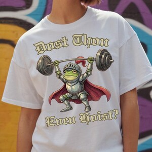 Pu&ograve; includere: T-shirt bianca con un ranocchio dei cartoni animati in armatura da cavaliere che solleva un bilanciere. Il ranocchio &egrave; verde con elmo e armatura argentati e un mantello rosso. Il testo "Dost Thou Even Hoist?" &egrave; stampato.