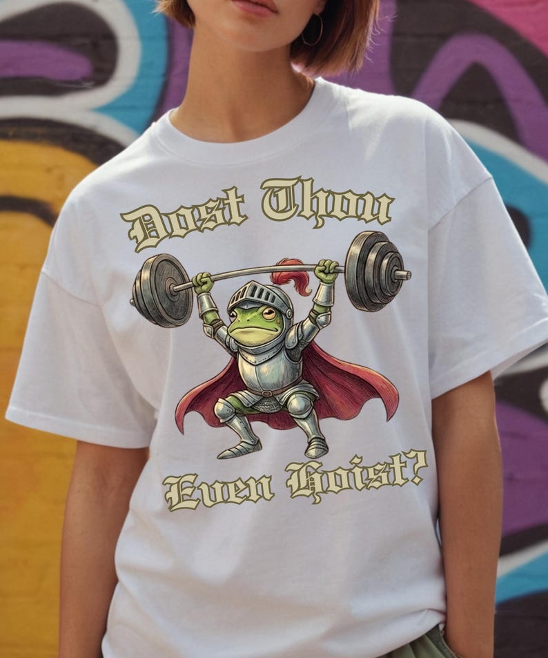 Pu&ograve; includere: T-shirt bianca con un ranocchio dei cartoni animati in armatura da cavaliere che solleva un bilanciere. Il ranocchio &egrave; verde con elmo e armatura argentati e un mantello rosso. Il testo "Dost Thou Even Hoist?" &egrave; stampato.