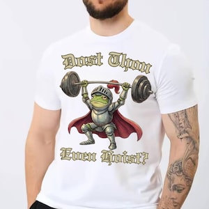 Pu&ograve; includere: T-shirt bianca con un ranocchio dei cartoni animati in armatura da cavaliere che solleva un bilanciere. La rana &egrave; verde con un mantello rosso e un elmo argentato. Il testo sulla maglietta recita "Dost Thou Even Hoist?"