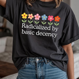 Könnte beinhalten: Dunkelgraues T-Shirt mit dem weißen Text "Radikalisiert durch grundlegende Anständigkeit". Oberhalb des Textes befindet sich eine Reihe bunter Blumen in Gelb, Pfirsich, Rosa, Blau, Rot und Orange.