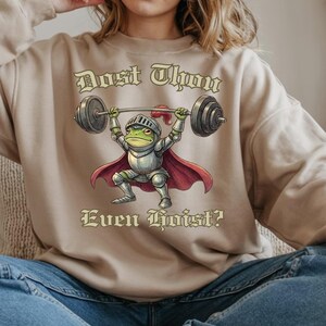 Pu&ograve; includere: Felpa beige con la scritta "Dost Thou Even Hoist?" e l'illustrazione di una rana in armatura da cavaliere che solleva un bilanciere. La rana &egrave; verde, con mantello e casco rossi.