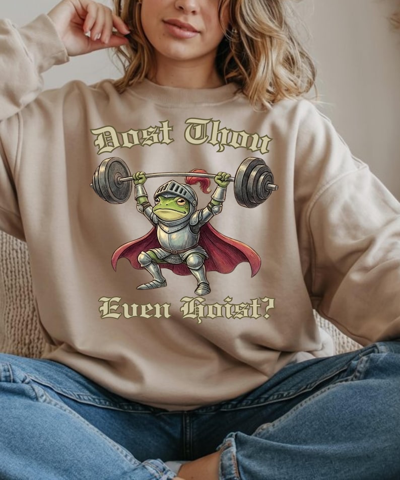 Pu&ograve; includere: Felpa beige con la scritta "Dost Thou Even Hoist?" e l'illustrazione di una rana in armatura da cavaliere che solleva un bilanciere. La rana &egrave; verde, con mantello e casco rossi.
