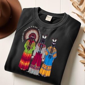 Könnte beinhalten: Schwarzes Sweatshirt mit Rundhalsausschnitt und einem farbenfrohen Motiv von drei indianischen Frauen in traditioneller Kleidung. Die Frauen sind mit langen Zöpfen und Kopfschmuck dargestellt und tragen Kleider in Rot, Grün und Blau. Das Sweatshirt hat ein Etikett mit der Aufschrift "Carr".