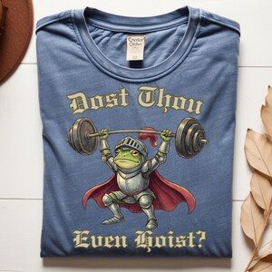 Pu&ograve; includere: T-shirt blu con un ranocchio dei cartoni animati in armatura da cavaliere che solleva pesi. Il ranocchio &egrave; verde con mantello e casco rossi. Il testo sulla maglietta recita "Dost Thou Even Hoist?"