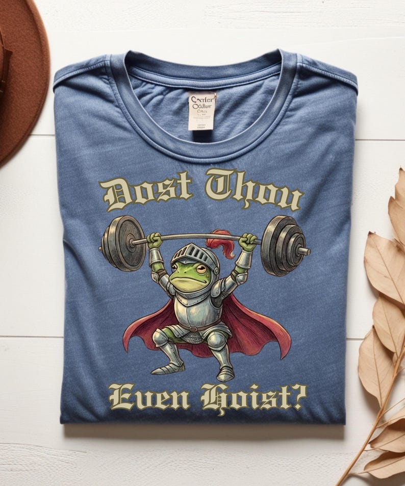 Pu&ograve; includere: T-shirt blu con un ranocchio dei cartoni animati in armatura da cavaliere che solleva pesi. Il ranocchio &egrave; verde con mantello e casco rossi. Il testo sulla maglietta recita "Dost Thou Even Hoist?"