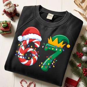 Op de afbeelding: Zwart sweatshirt met een feestelijk kerstontwerp. Het cijfer 6 is gestileerd als een zuurstok met een kerstmuts, en het cijfer 7 is een elfenmuts. Beide cijfers zijn versierd met kleurrijke kerstverlichting.