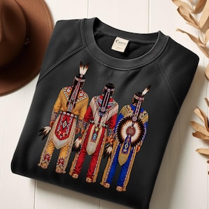Peut inclure: Un sweat-shirt noir à manches longues orné d'une impression graphique représentant trois personnages vus de dos, portant des tenues traditionnelles détaillées et colorées avec des plumes, des franges et des motifs géométriques.
