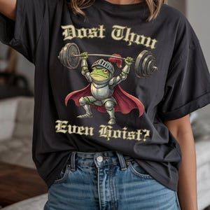 Pu&ograve; includere: T-shirt grigio scuro con un ranocchio dei cartoni animati in armatura da cavaliere che solleva un bilanciere. La rana indossa un elmo e un mantello rosso, con il testo "Dost Thou Even Hoist?" in un carattere ornato.