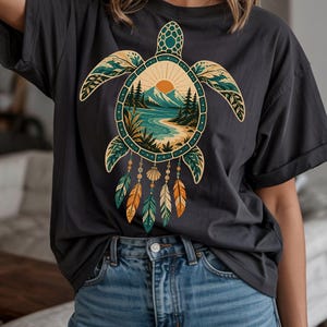 May include: A charcoal gray t-shirt featuring a large graphic of a sea turtle. The turtle shell contains a landscape illustration of a sun, mountains, and a beach. Dangling from the bottom of the shell are several feathers and a seashell.