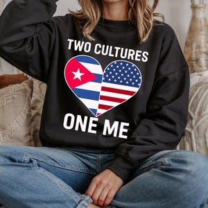 May include: Black sweatshirt with the text "TWO CULTURES ONE ME" in white. A heart-shaped graphic features the Cuban and American flags. The sweatshirt is worn with blue jeans.