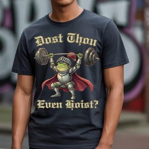 Pu&ograve; includere: T-shirt blu navy scuro con un ranocchio dei cartoni animati in armatura da cavaliere che solleva un bilanciere. Il testo "Dost Thou Even Hoist?" &egrave; stampato sopra e sotto l'illustrazione della rana in oro.