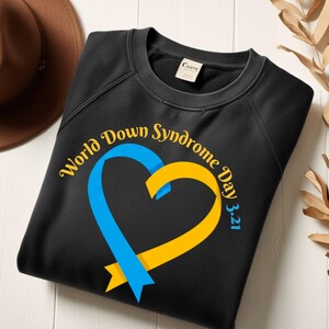 Peut inclure: Sweat-shirt noir avec un cœur en ruban bleu et jaune. Le texte "World Down Syndrome Day 3.21" est disposé en arc de cercle autour du cœur. Un chapeau marron et des feuilles séchées sont en arrière-plan.