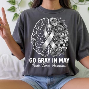 Könnte beinhalten: Dunkelgraues T-Shirt mit einem grafischen Design, das ein Gehirn, Blumen und ein silbernes Band zeigt. Der Text "GO GRAY IN MAY Brain Tumor Awareness" ist darunter gedruckt. Das Shirt ist ein lässiger Stil.