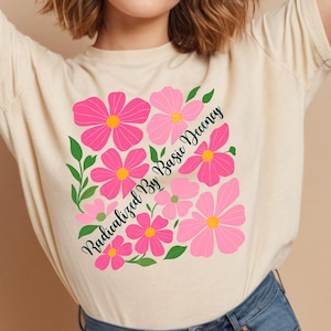 Può includere: T-shirt color crema con un design floreale di fiori rosa e foglie verdi. Il testo "Radicalized By Basic Decency" è stampato in diagonale in nero. La maglietta è realizzata in un materiale morbido.
