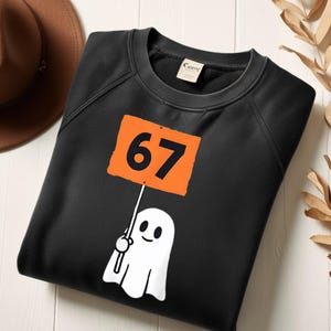 May include: Black sweatshirt featuring a white ghost holding a sign with the number 67 in black on an orange background. The sweatshirt has a crew neck and long sleeves. A brown hat is visible in the upper left corner.