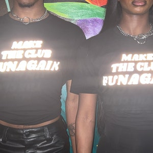 May include: Two people wearing black t-shirts with reflective white text that reads "Make the club fun again".