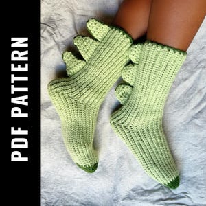 Puede incluir: Un par de calcetines de dinosaurio de crochet verde con ribete verde y marrón. Los calcetines tienen un diseño de pie de dinosaurio en la parte superior.