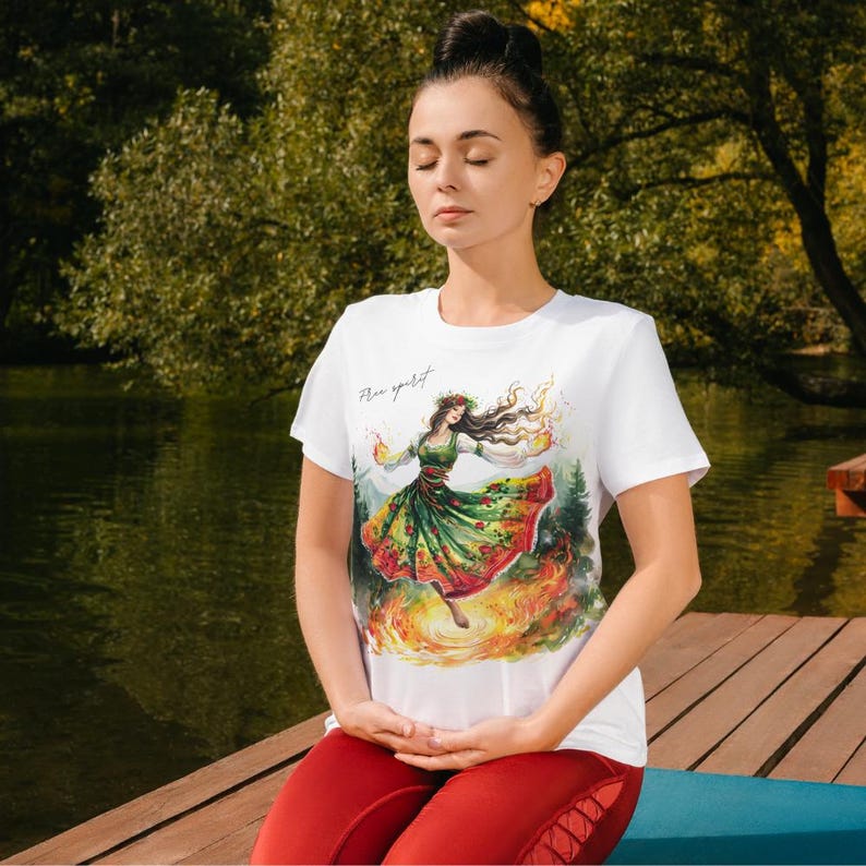 Puede incluir: Camiseta blanca con una ilustraci&oacute;n colorida de una mujer bailando en un bosque con llamas a su alrededor. El texto "Fire spirit" est&aacute; impreso en la camiseta.