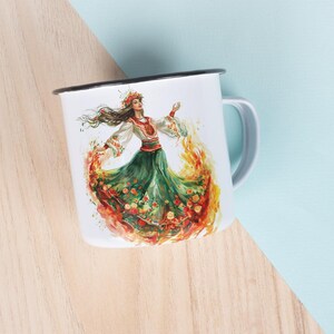 Puede incluir: Taza de esmalte blanco con una ilustraci&oacute;n colorida de una mujer con un vestido verde y rojo bailando con llamas a su alrededor. La mujer lleva una corona de flores y una blusa blanca bordada.