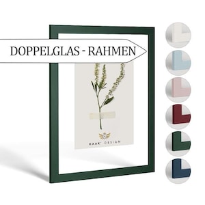 Op de afbeelding: Een donkergroen fotolijst met een witte passe-partout en een geperste bloem erin. De lijst heeft de tekst "Doppelglas - Rahmen" en "HAAK DESIGN" erop gedrukt.