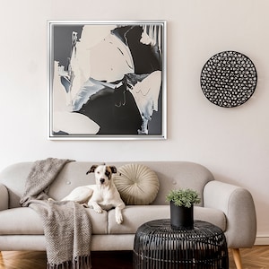 May include: A white dog with black and brown spots is lying on a light gray couch with a brown throw blanket. There is a black and white abstract painting on the wall behind the couch. A black wicker side table is in front of the couch.