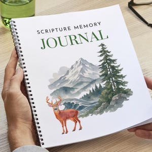 Può includere: Un diario con rilegatura a spirale con copertina bianca che riporta il testo SCRIPTURE MEMORY JOURNAL in nero e verde. La copertina presenta un'illustrazione ad acquerello di un cervo marrone davanti a una catena montuosa e un pino.