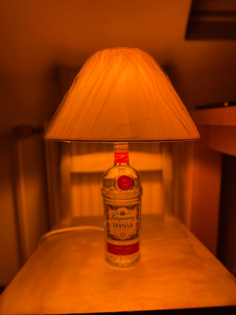 May include: A distinctive table lamp crafted from a Tanqueray Sevilla gin bottle. The clear glass bottle supports a cream-coloured lampshade. The lamp is positioned on a wooden chair, producing a warm, inviting atmosphere.