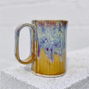 May include: A ceramic mug featuring a tall, rectangular handle and a cylindrical body. The glaze transitions from a deep mustard yellow at the base to a mottled blend of light blue, white, and purple drips cascading down from the rim.