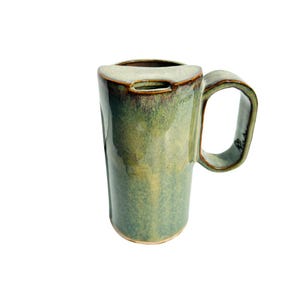 May include: A tall, green ceramic mug with a unique handle and a built-in lid. The mug has a glossy finish with variations in the green color. The handle is rectangular and the rim has a darker brown accent. The mug is designed for hot beverages.