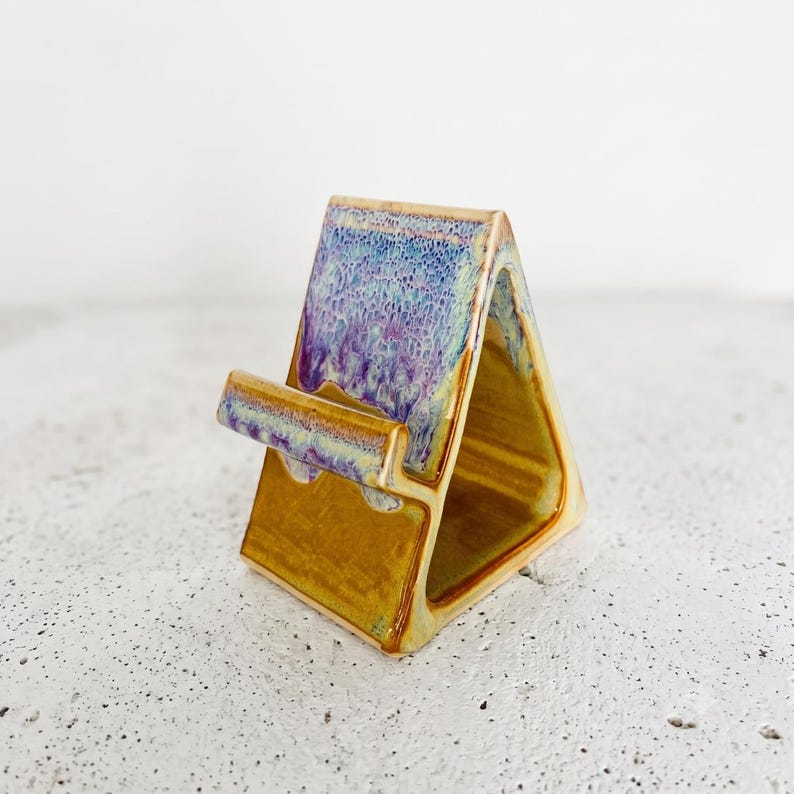 May include: A triangular ceramic phone stand featuring a glossy finish. The top section displays a textured, mottled glaze in shades of blue and purple, while the base and interior are finished in a smooth, solid mustard yellow glaze.